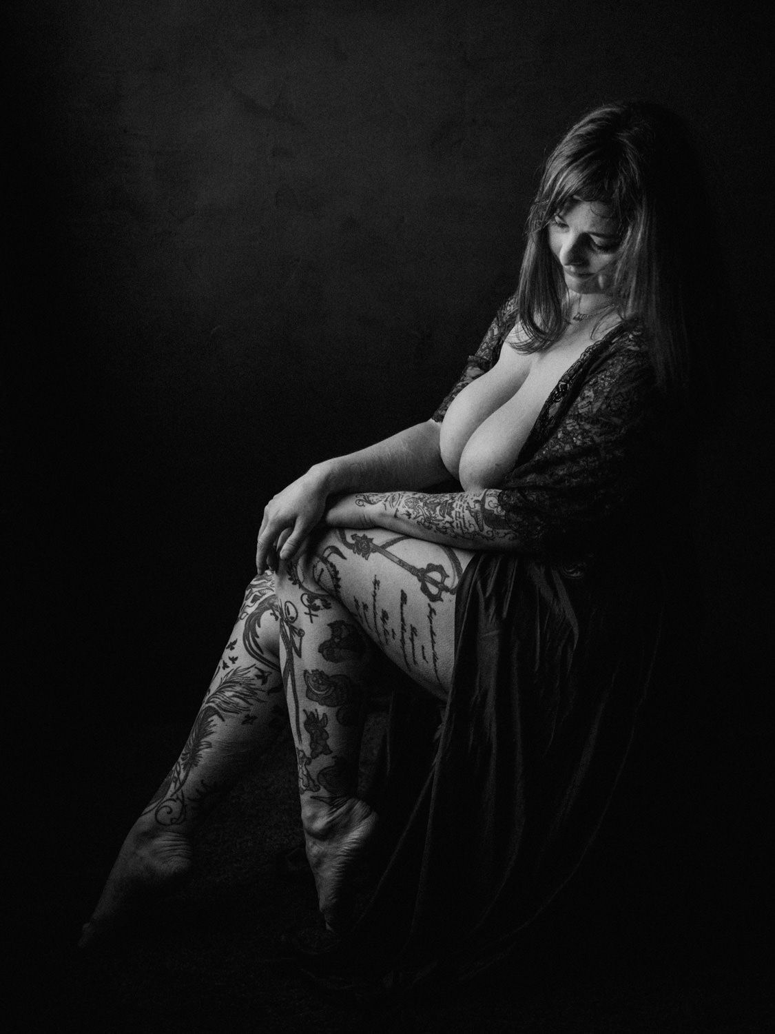 Black and white fine art portrait of a tattooed woman in a dark studio setting.