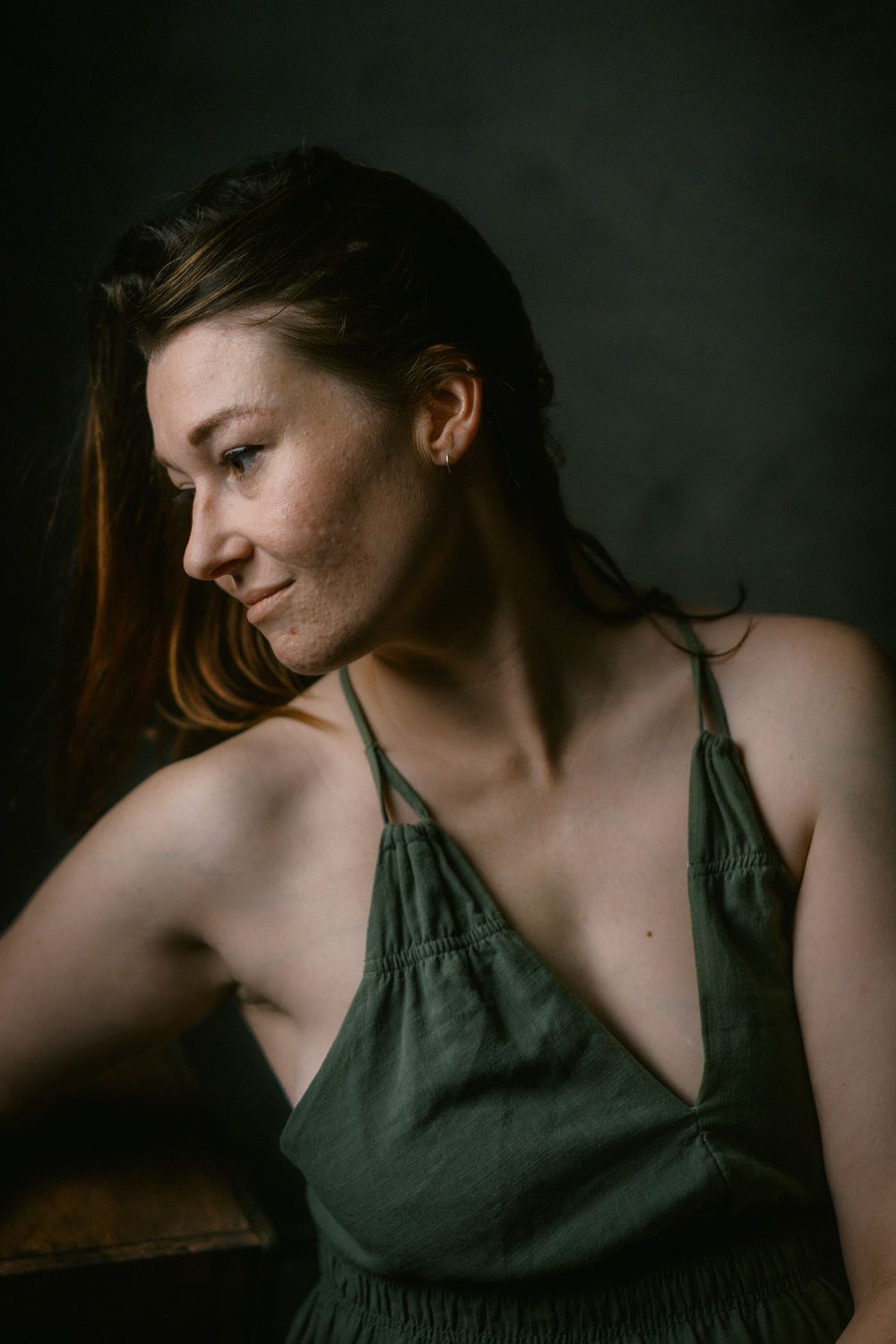 A young woman in a green linen halter dress gazes downward against a dark moody studio background.