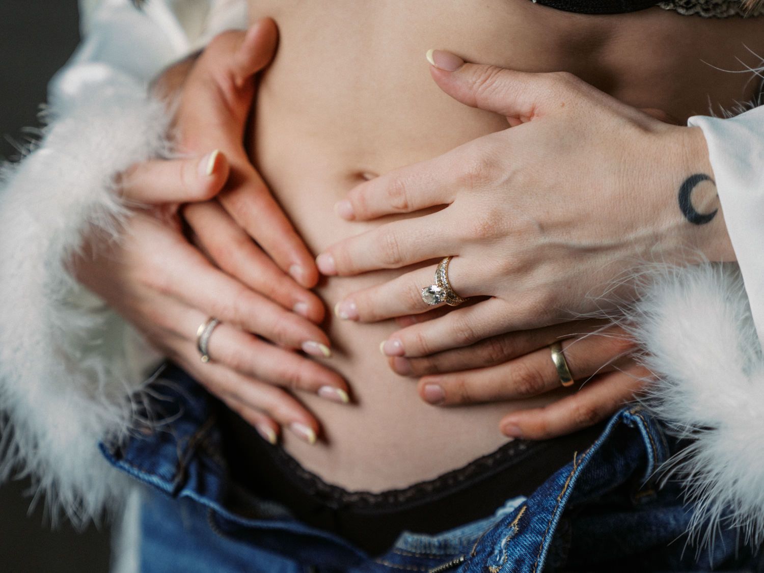 Two pairs of hands gently cradling a pregnant woman's bare belly, wearing rings, over blue jeans.