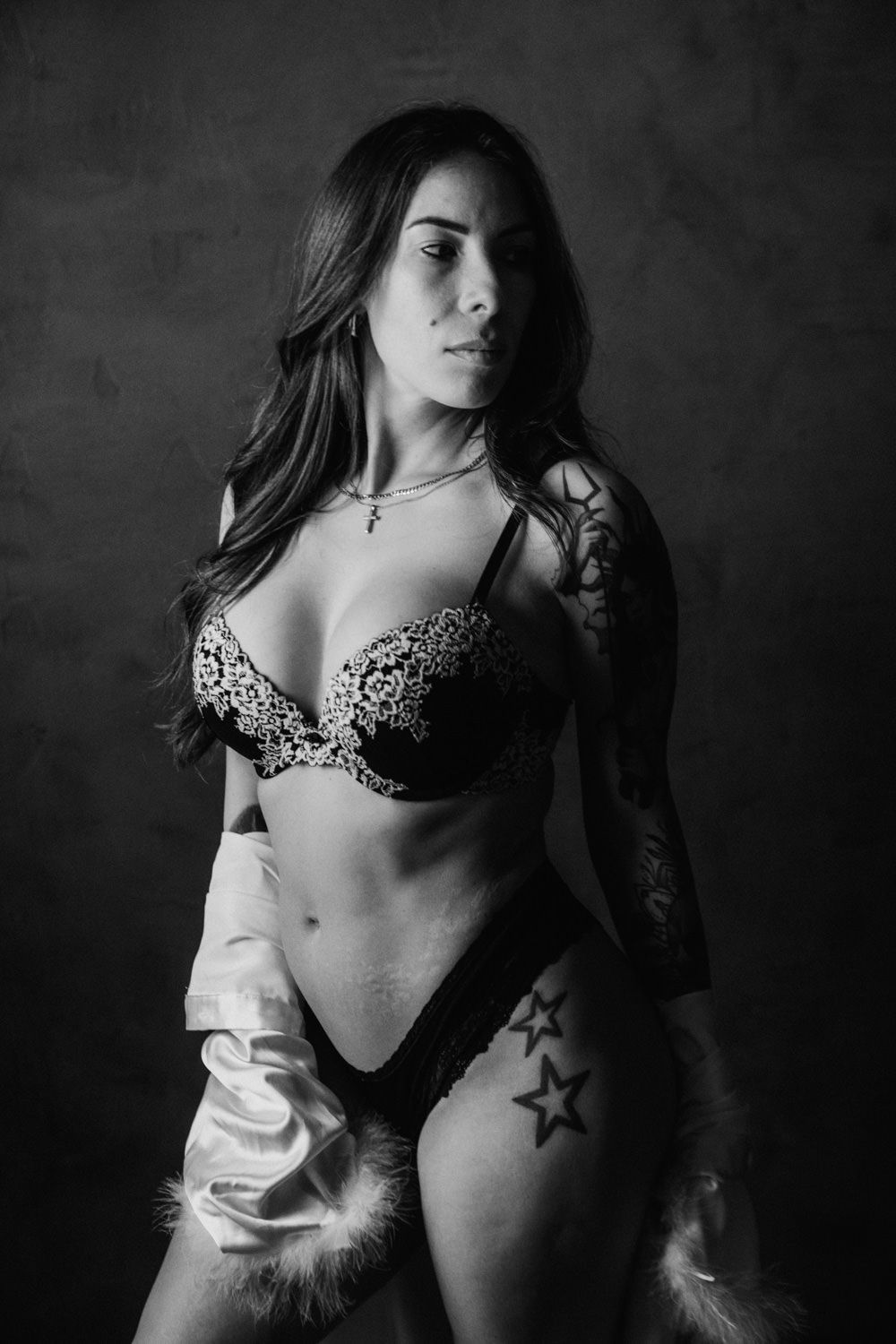 Black and white boudoir photo of a woman in lace lingerie with star tattoos, posing in a satin robe against dark backdrop.