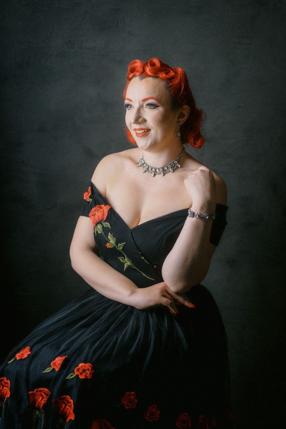 Woman in black floral off-shoulder dress with red hair, diamond necklace, posing elegantly against dark background.