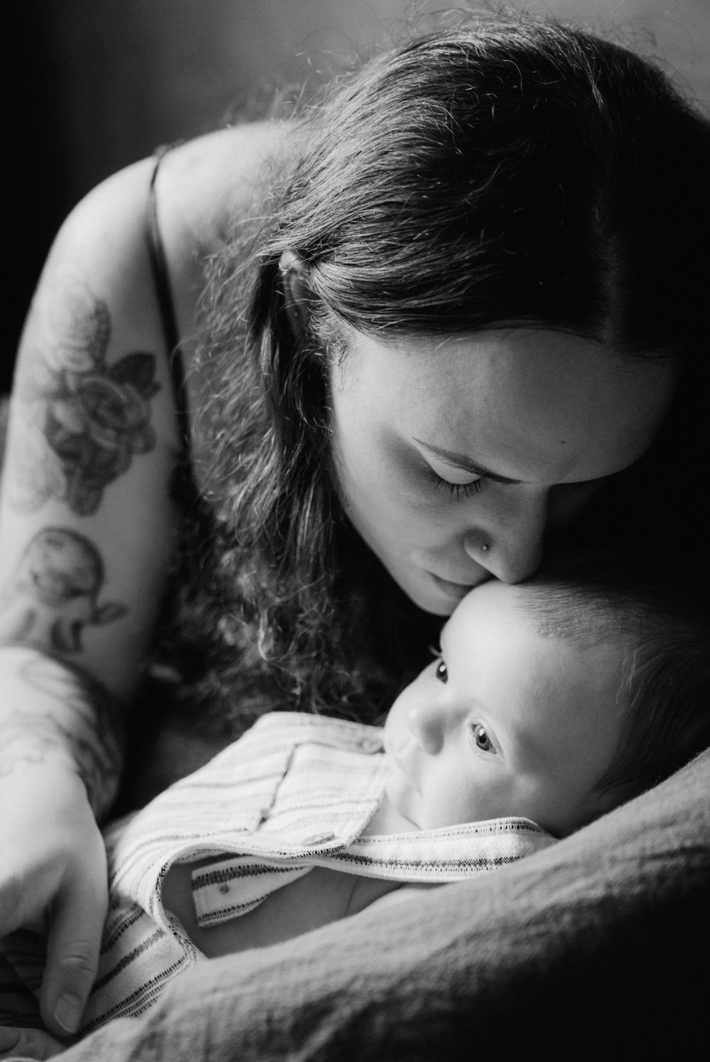 Black and white photo of a tattooed mother tenderly nuzzling her newborn baby while cradling them gently.