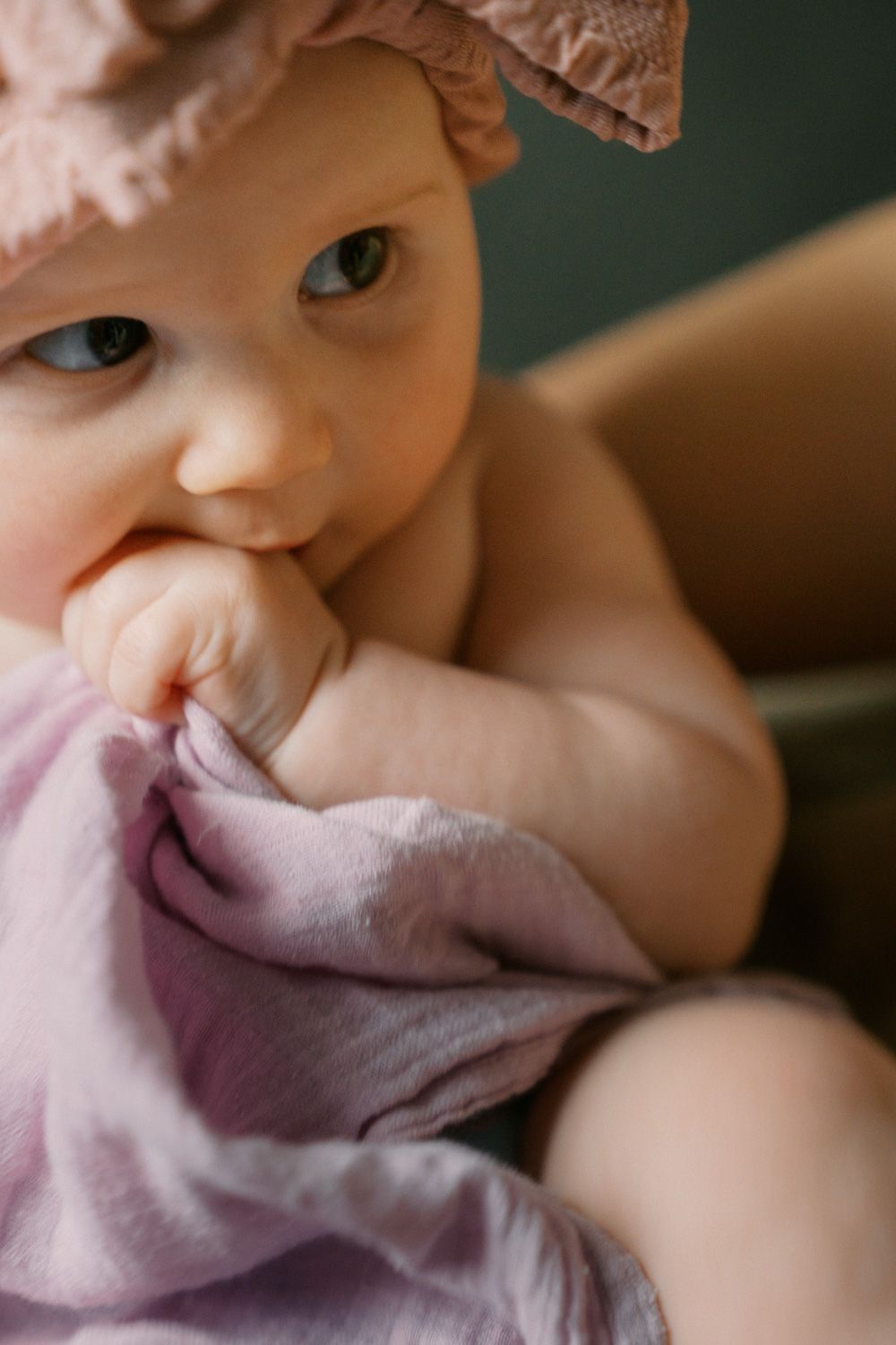 Adorable baby girl in pink hat chewing on a soft lavender blanket, looking curiously at the camera.