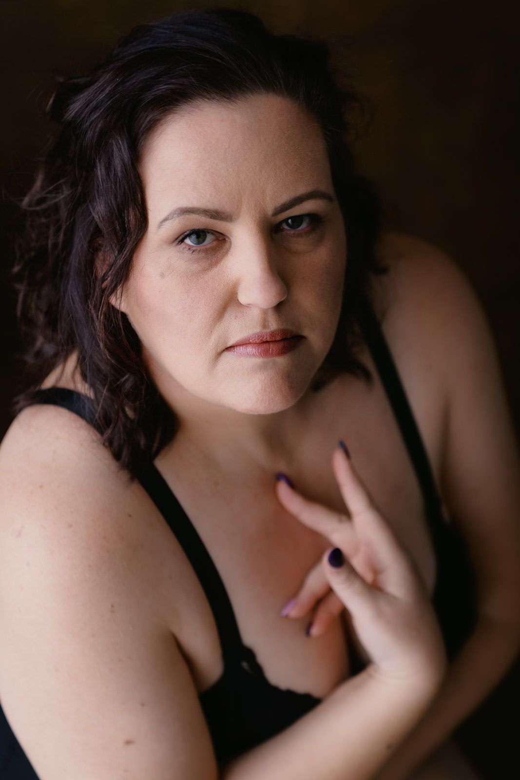 A woman with dark curly hair in black lingerie poses confidently against a dark background.