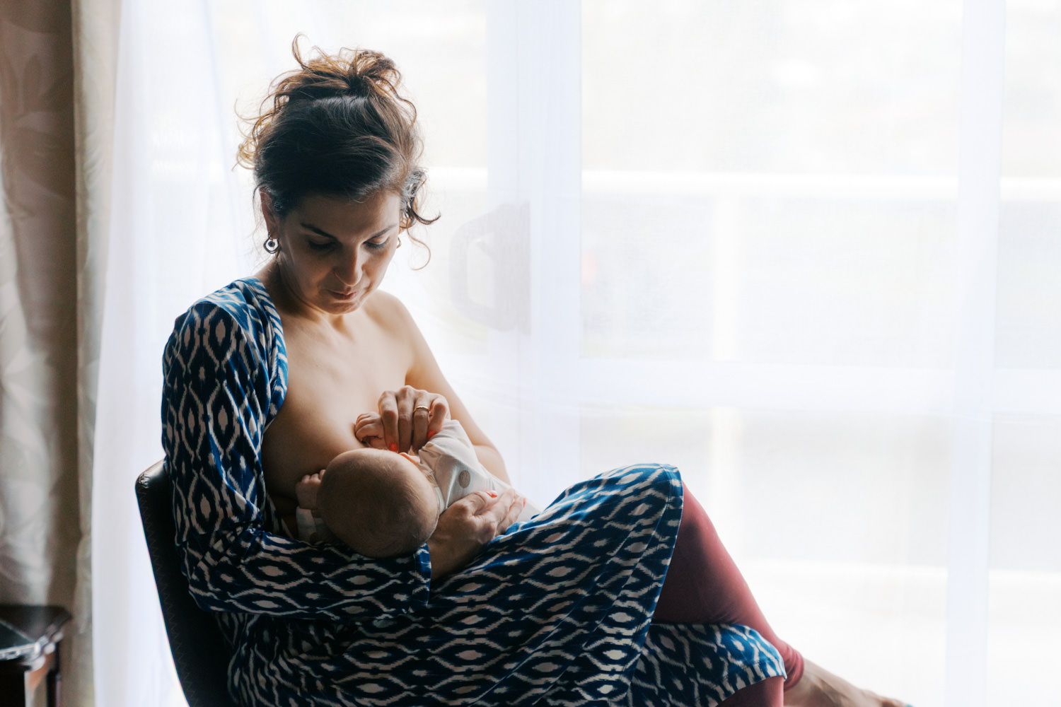 Mother breastfeeding newborn baby while sitting in armchair near window wearing blue patterned robe.