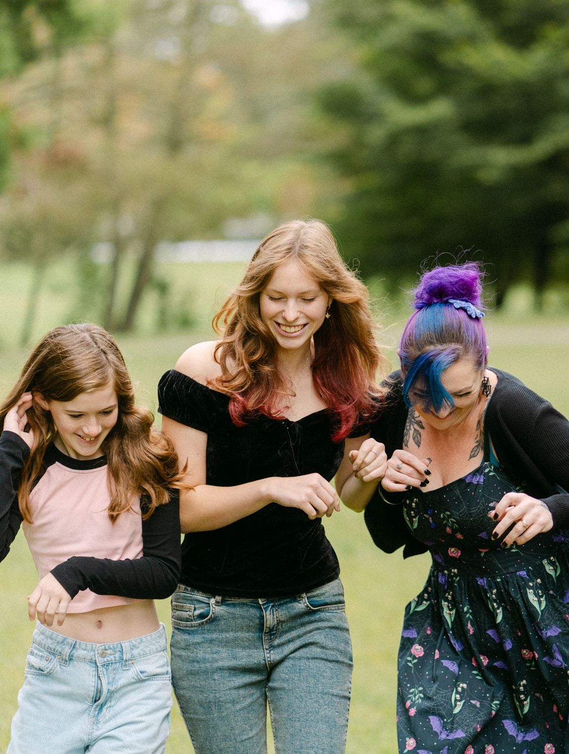 Three laughing teenage girls with colorful hair walk together outdoors in a lush green park setting.