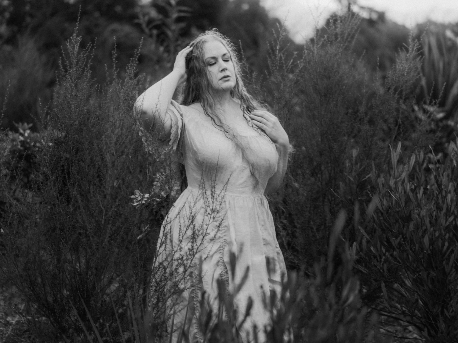 Black and white photo of a woman in a lace dress standing among lush green plants, hands in her hair.