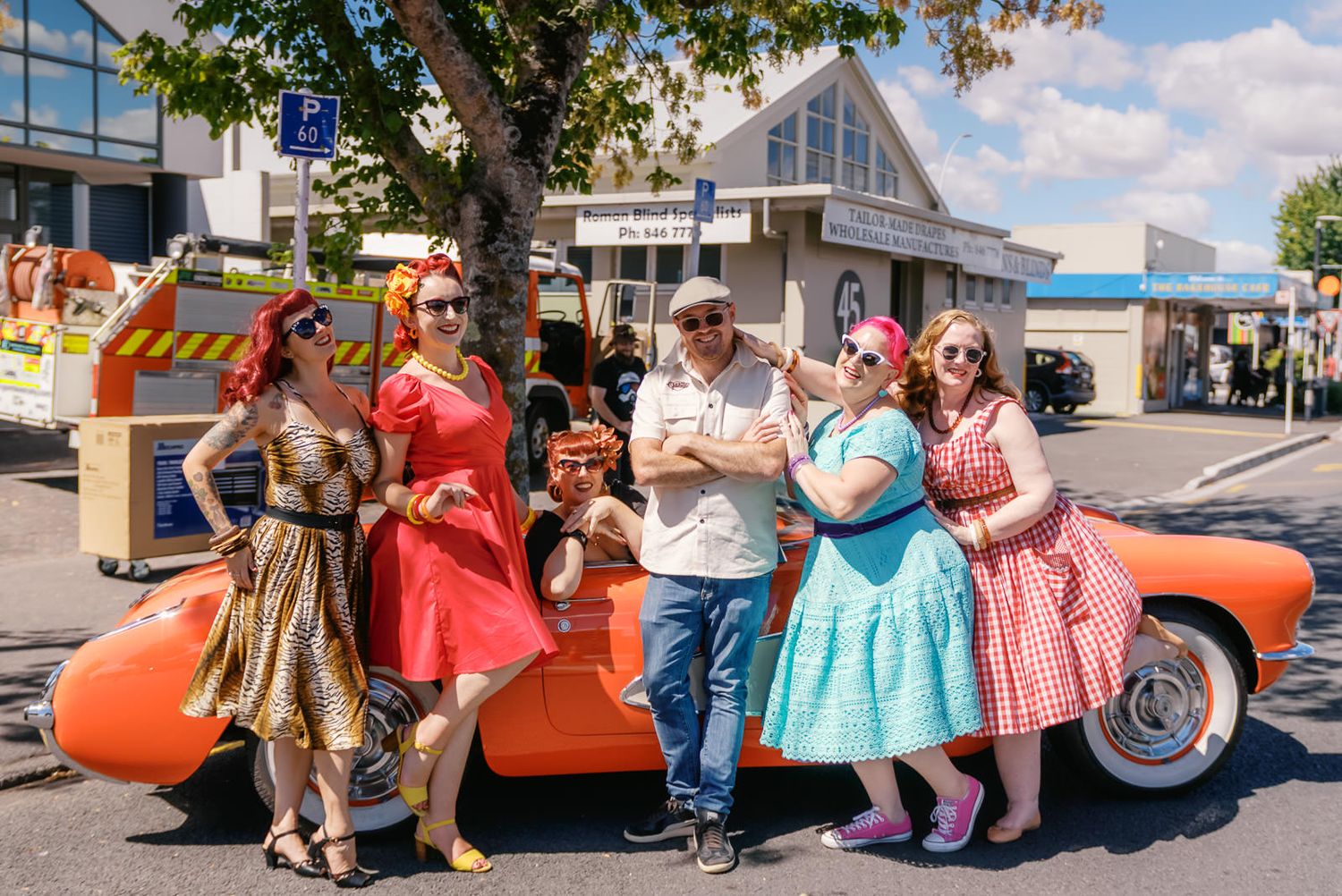 Group of people in 1950s retro outfits posing beside a classic orange car at a sunny outdoor event.