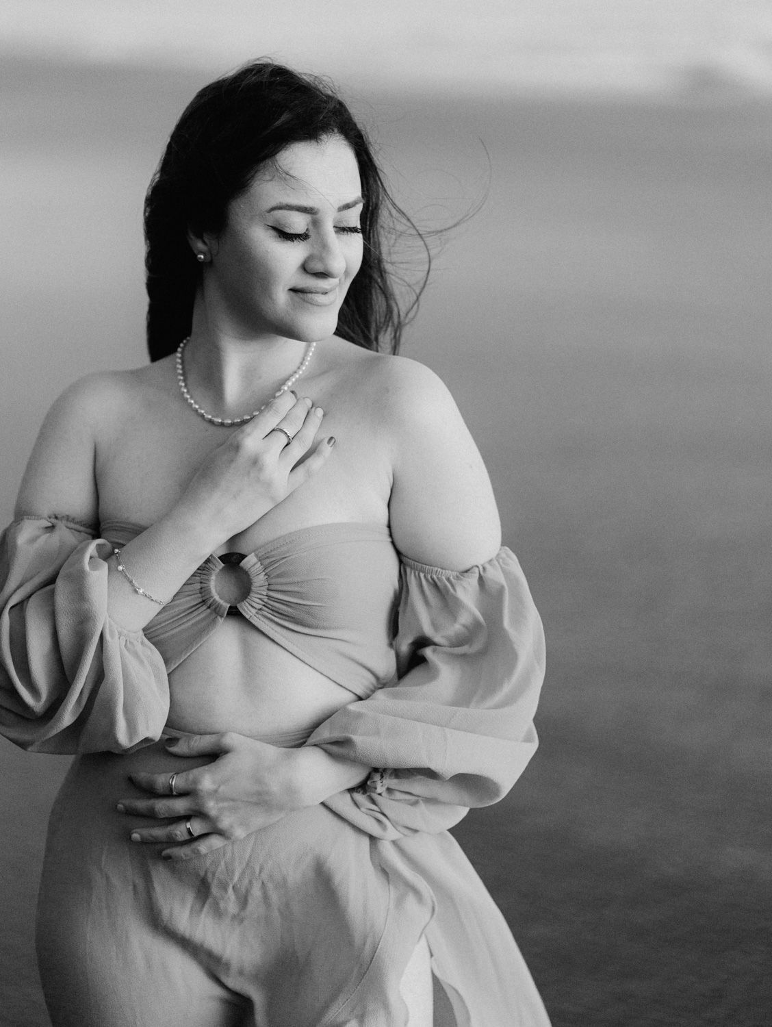Black and white maternity photo of a woman in a flowing off-shoulder dress, posing gracefully outdoors.