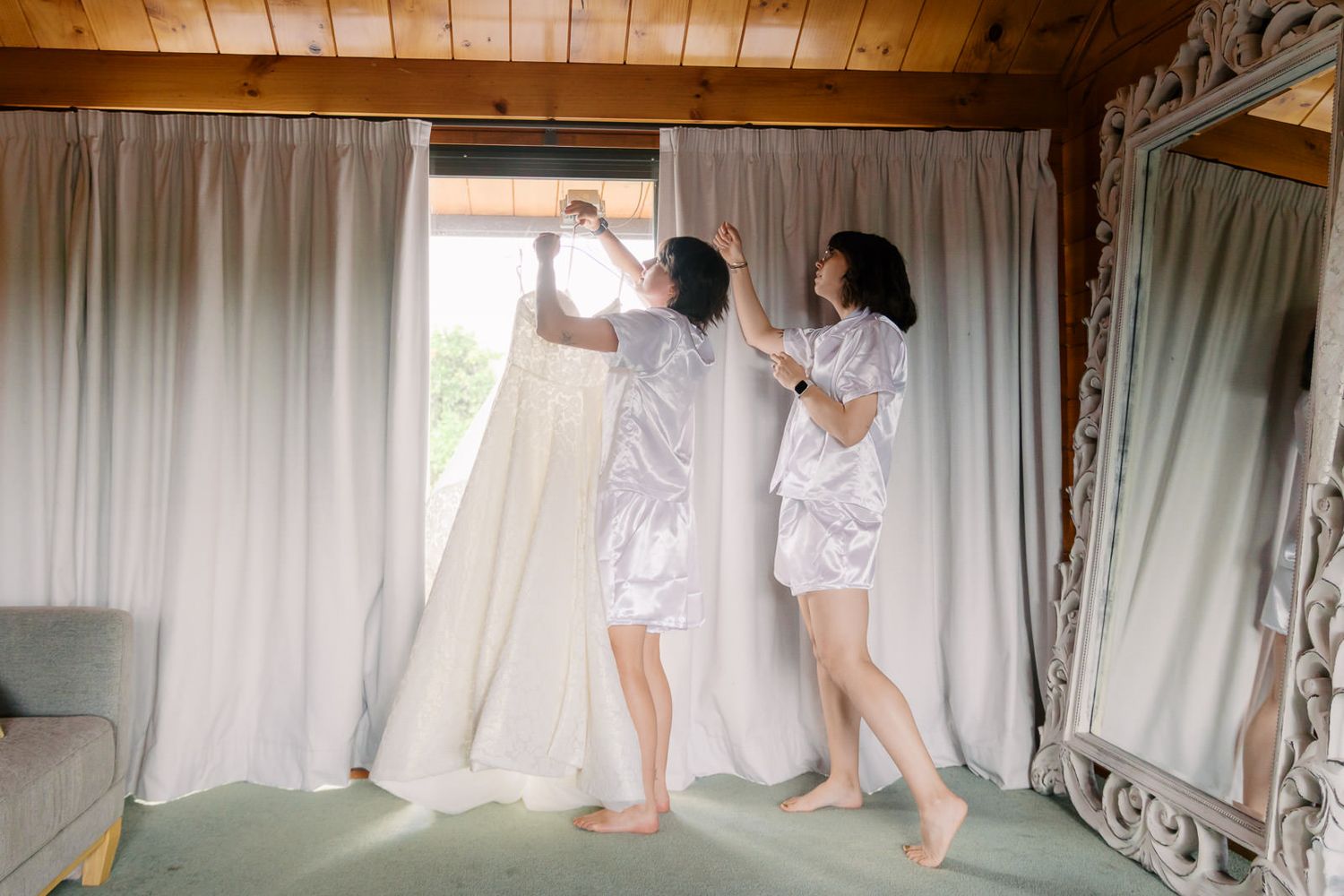 Two bridesmaids in white satin robes lifting a wedding dress near sheer curtains in an elegant rustic venue.