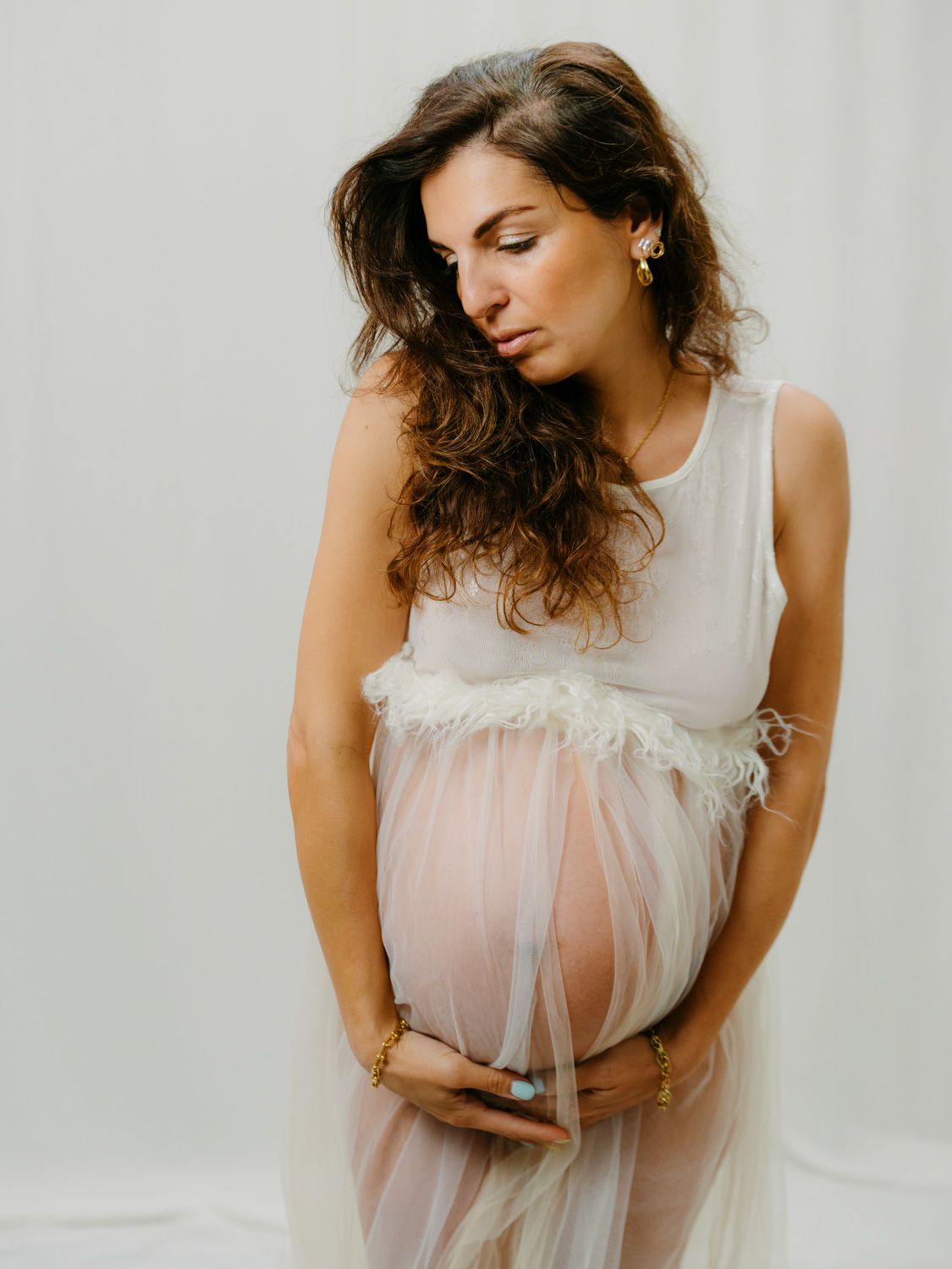 Pregnant woman in sheer white maternity gown cradling her baby bump during an elegant studio photoshoot.