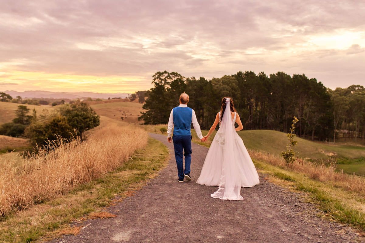 Bride and groom walk hand-in-hand down a rural path at sunset, surrounded by golden fields and rolling hills.