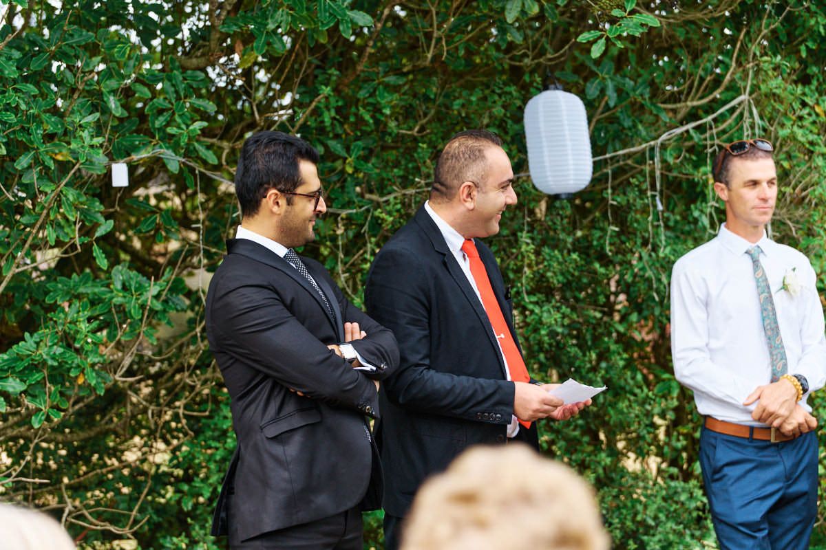Two men in suits stand outdoors at a wedding ceremony, one holding papers, surrounded by lush green foliage.