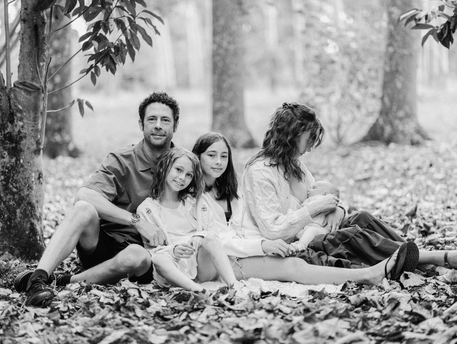 Black and white family portrait of parents and two children sitting together among fallen leaves in a forest.