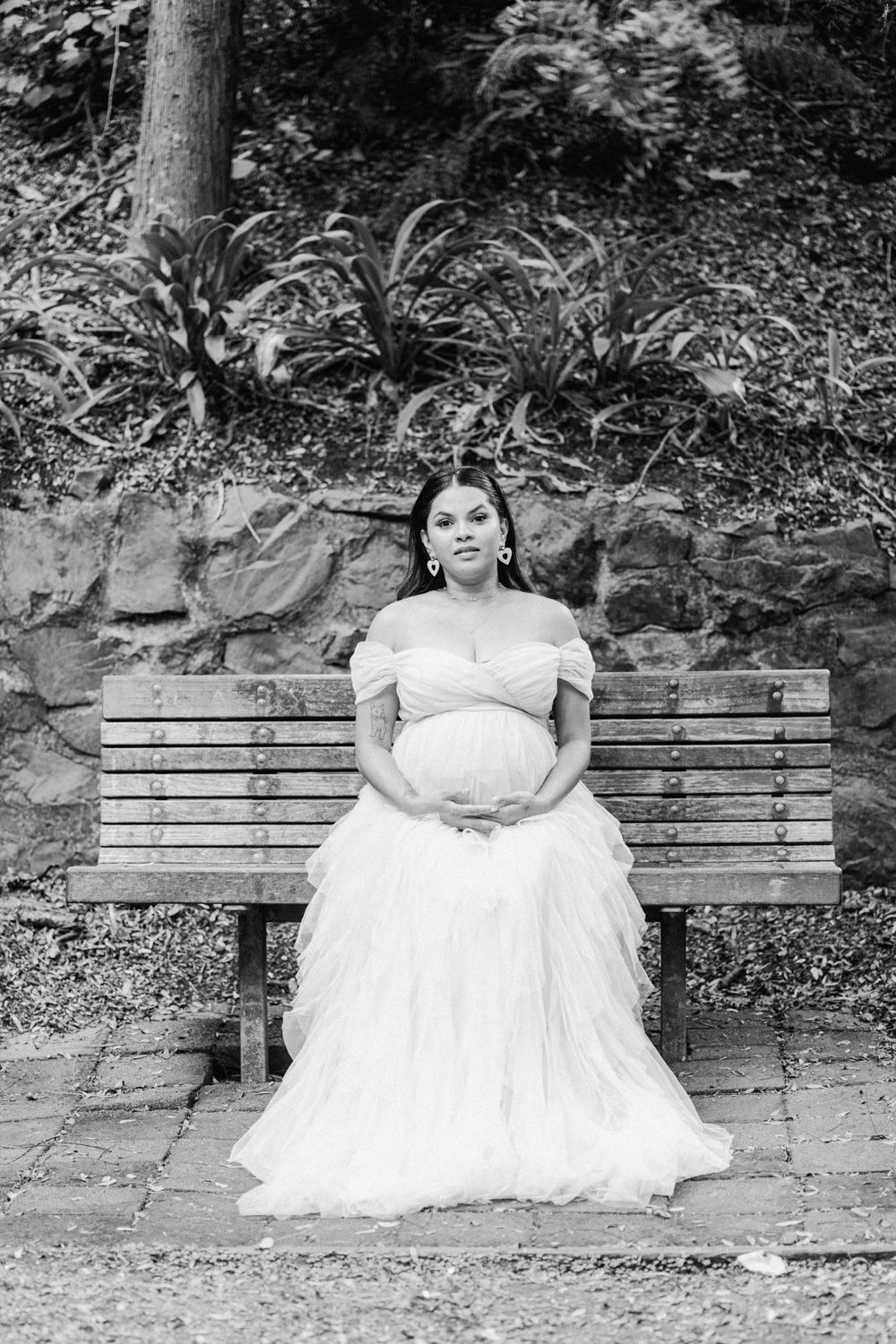 Pregnant woman in white off-shoulder gown sits on wooden bench in garden for elegant maternity portrait.