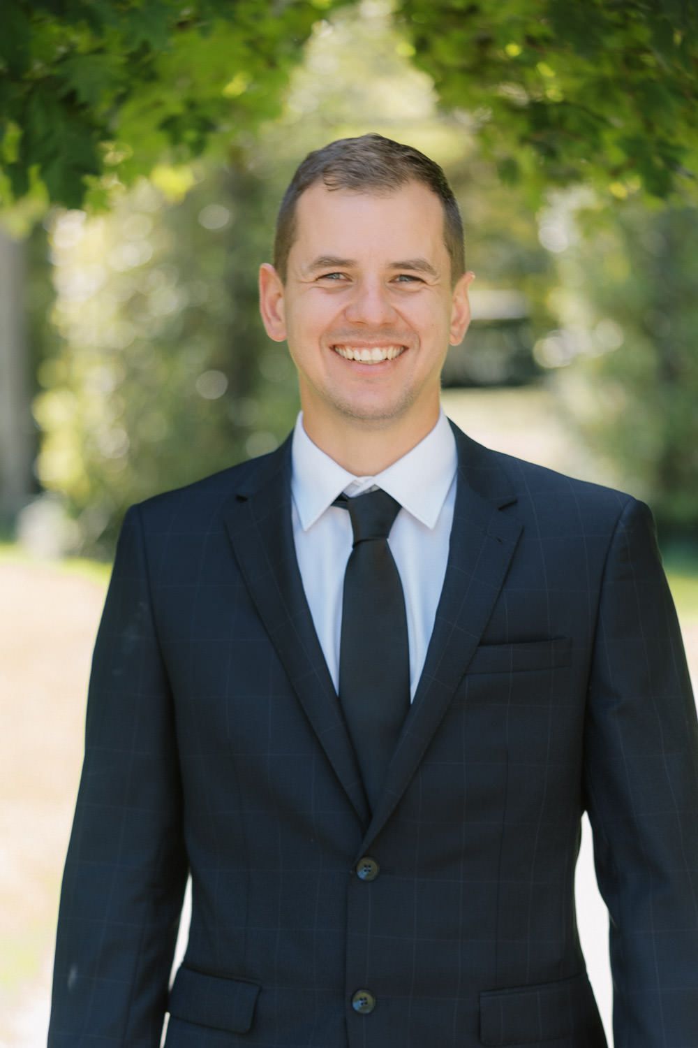 Smiling businessman in dark suit and black tie standing outdoors among green trees on a sunny day.