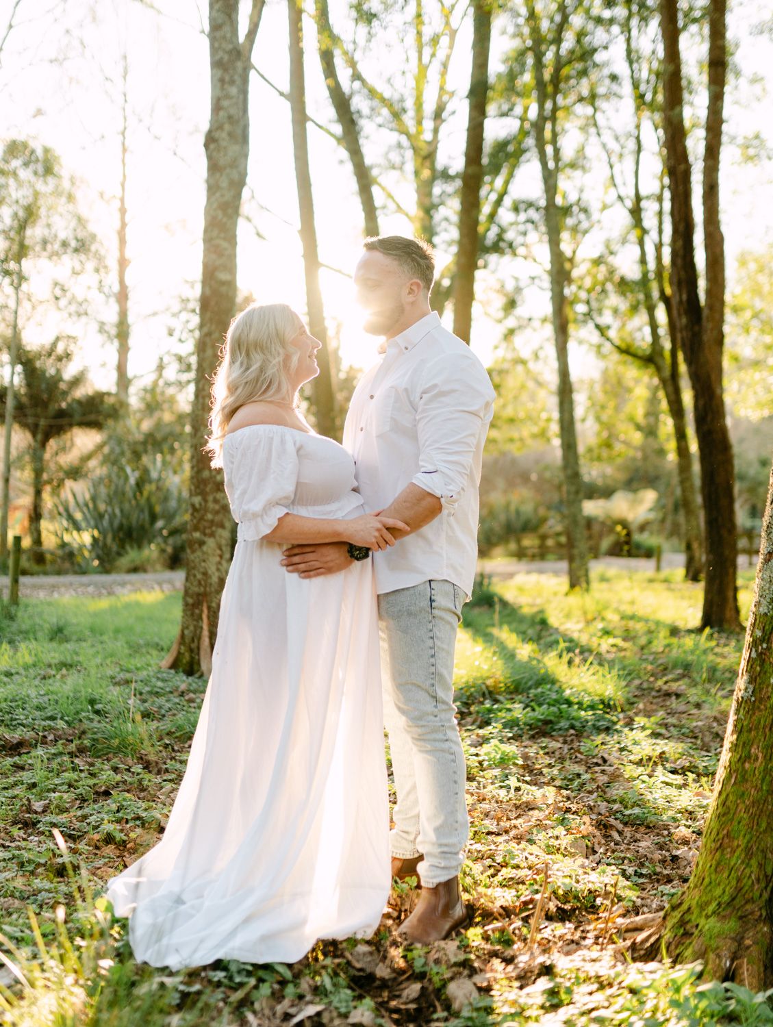 Couple embracing in sunlit forest, woman in flowing white maternity gown, man in white shirt and grey jeans.