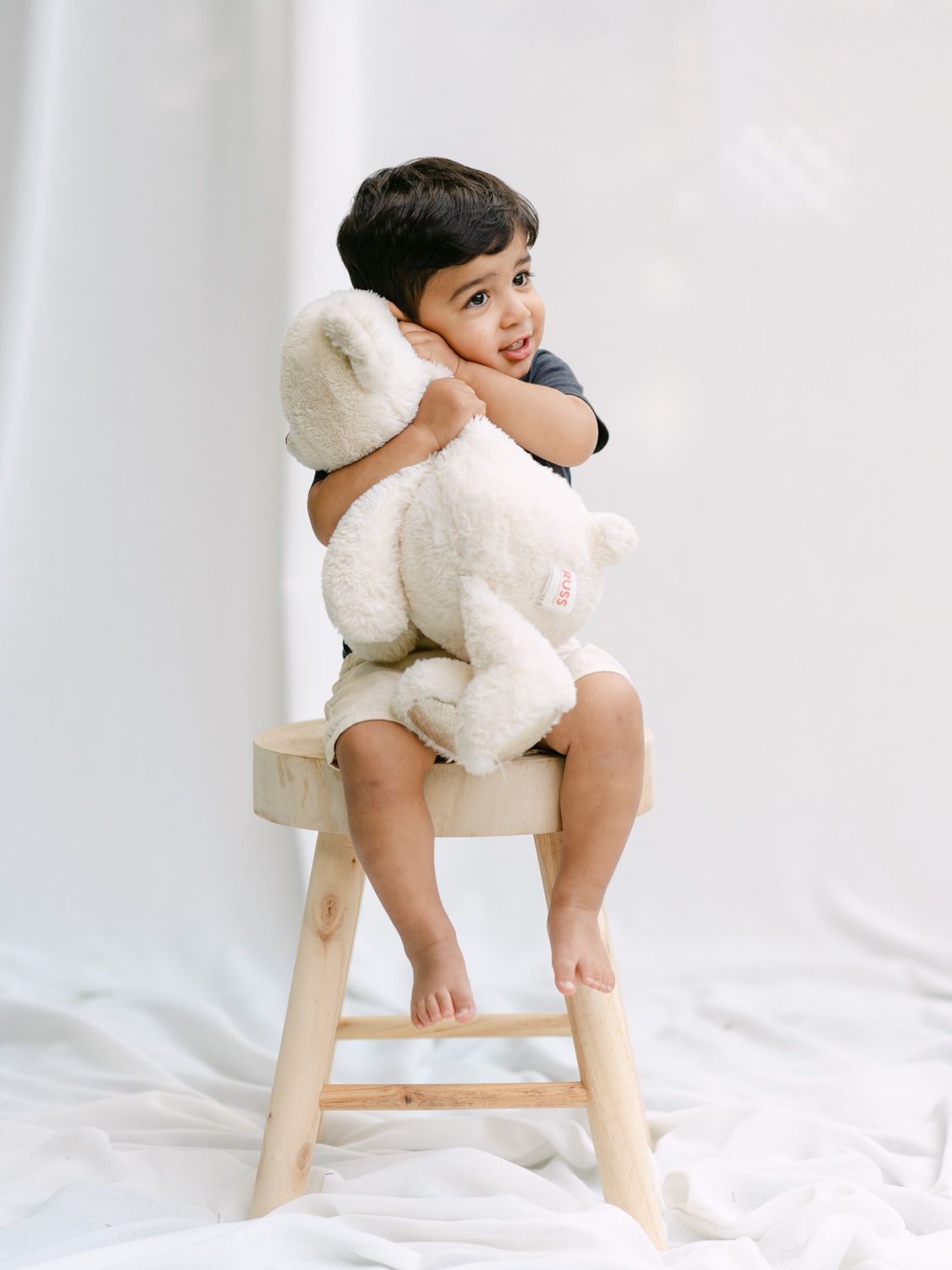 A toddler sits on a wooden stool hugging a white teddy bear, smiling over their shoulder against a white background.