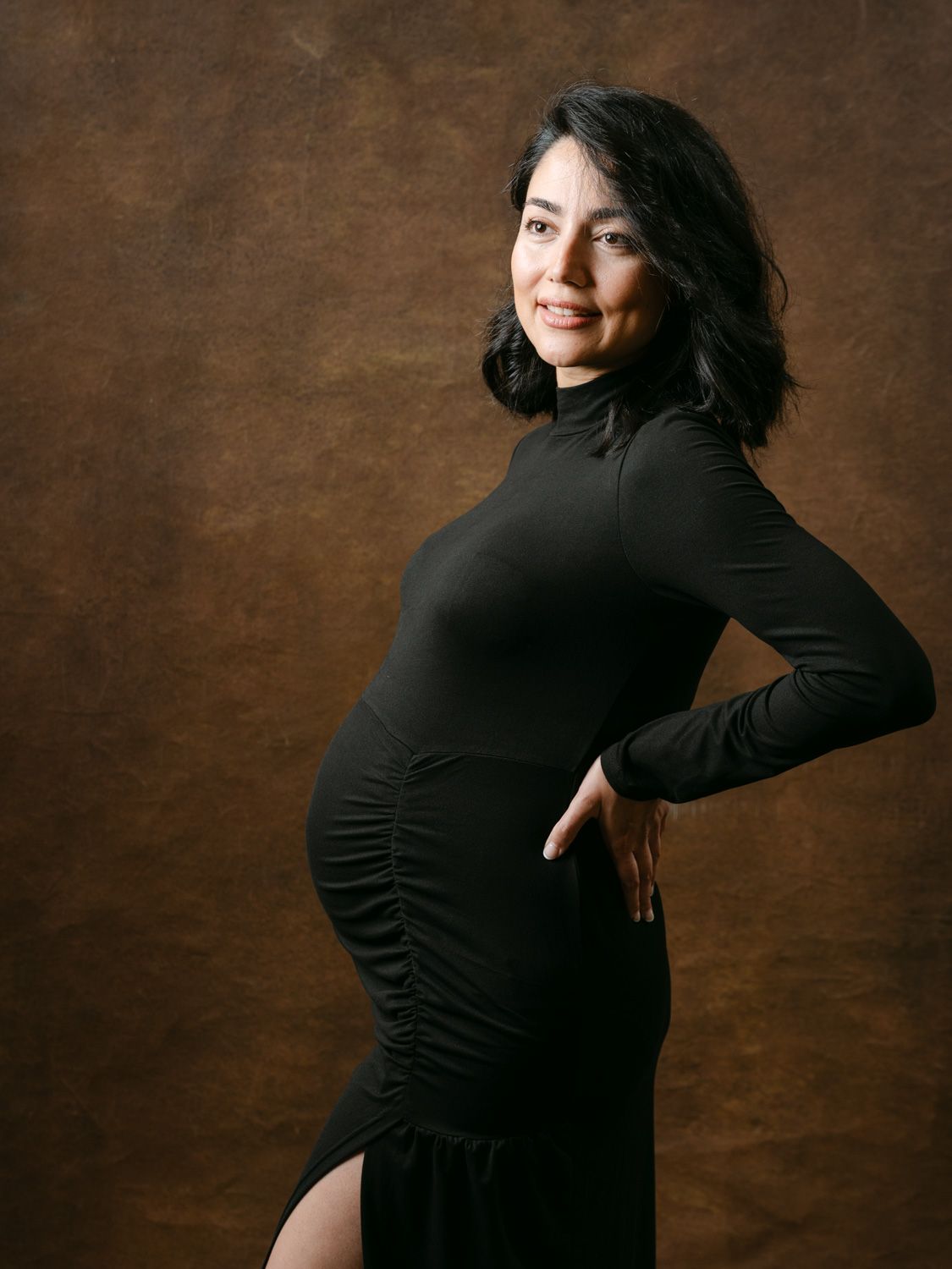 Pregnant woman in black turtleneck dress posing sideways, smiling, against brown studio backdrop.