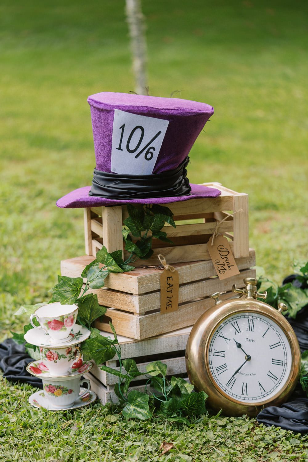 Alice in Wonderland Mad Hatter tea party display with purple top hat, vintage clock, teacups, and ivy on wooden crate.