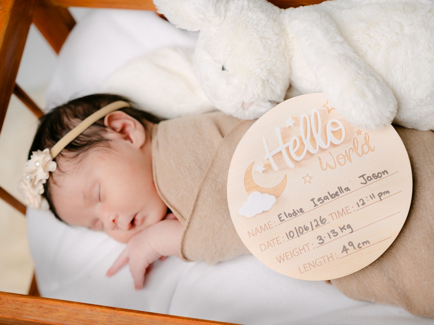 Newborn baby girl sleeping peacefully in crib with white bunny plush and Hello World birth announcement plaque.