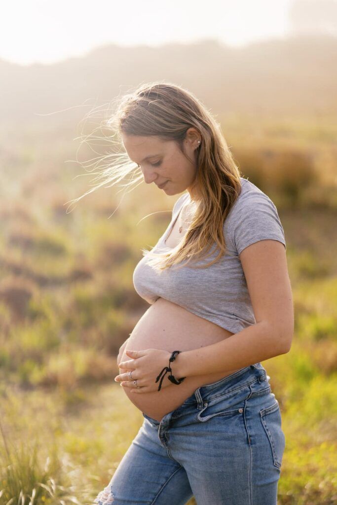 Pregnant woman in gray top and jeans cradling her baby bump outdoors in a sunny field with wind-blown hair.