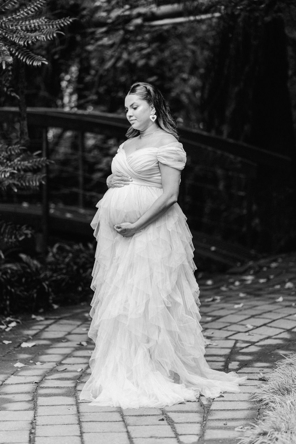 Black and white maternity photo of a pregnant woman in a ruffled white gown, cradling her belly in a lush garden.