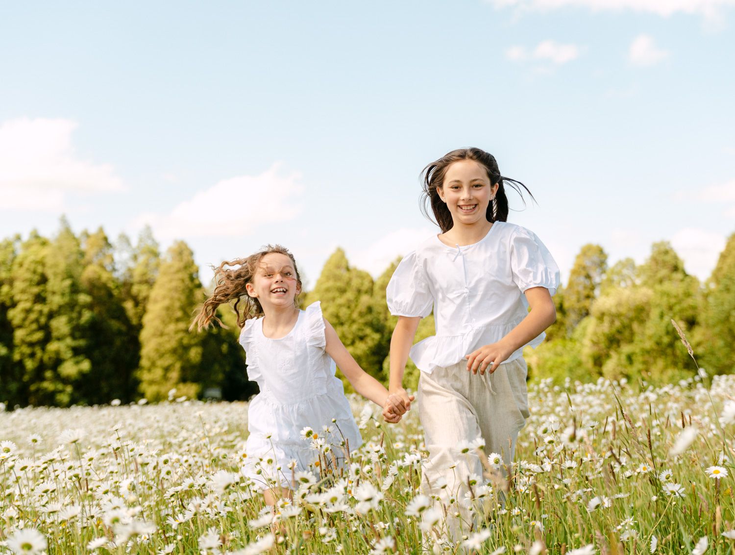 Two girls in white dresses running through a daisy field with trees and blue sky in the background.