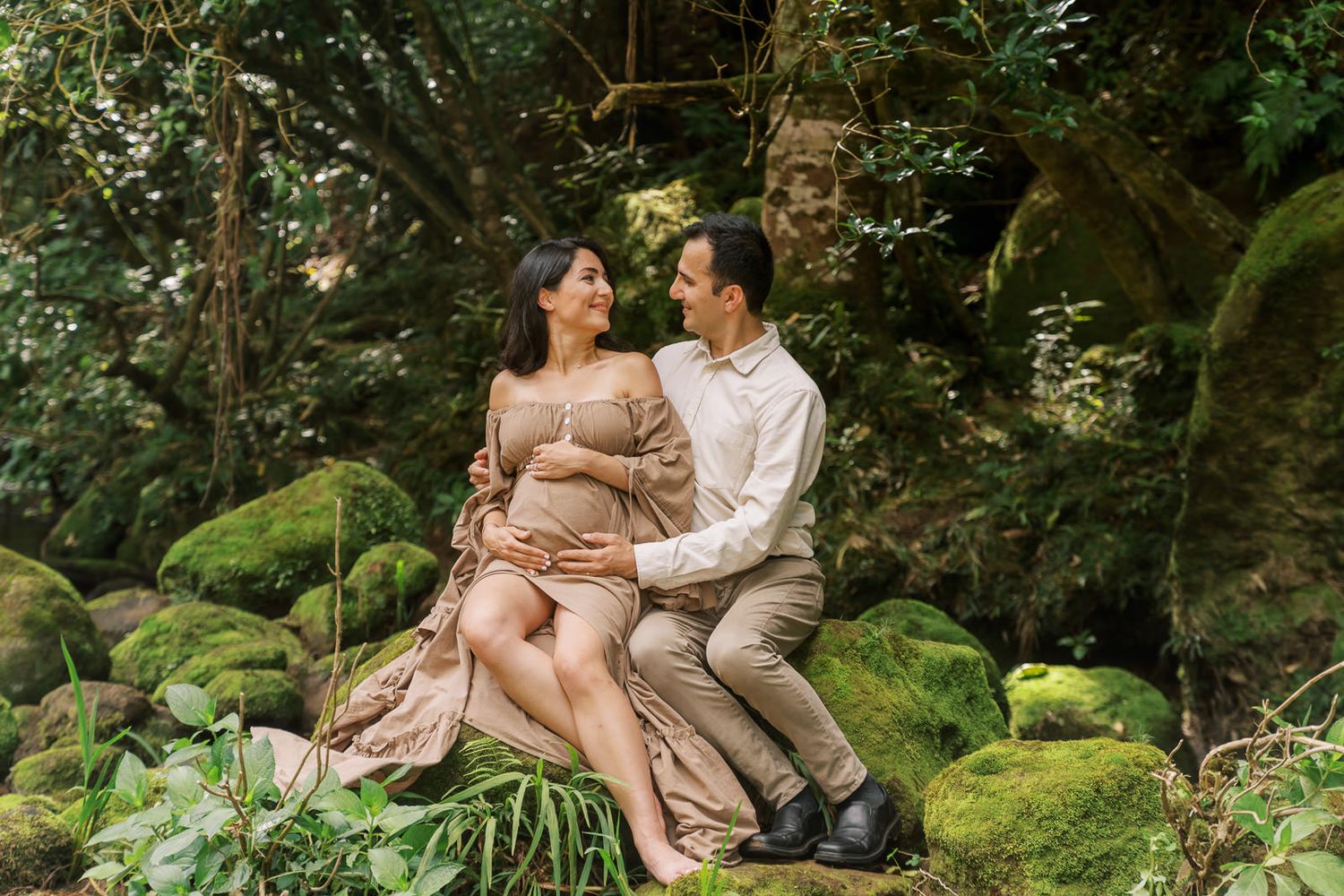 Couple poses on mossy rocks in lush forest, woman in flowing maternity dress, man in beige shirt, sharing a tender moment.