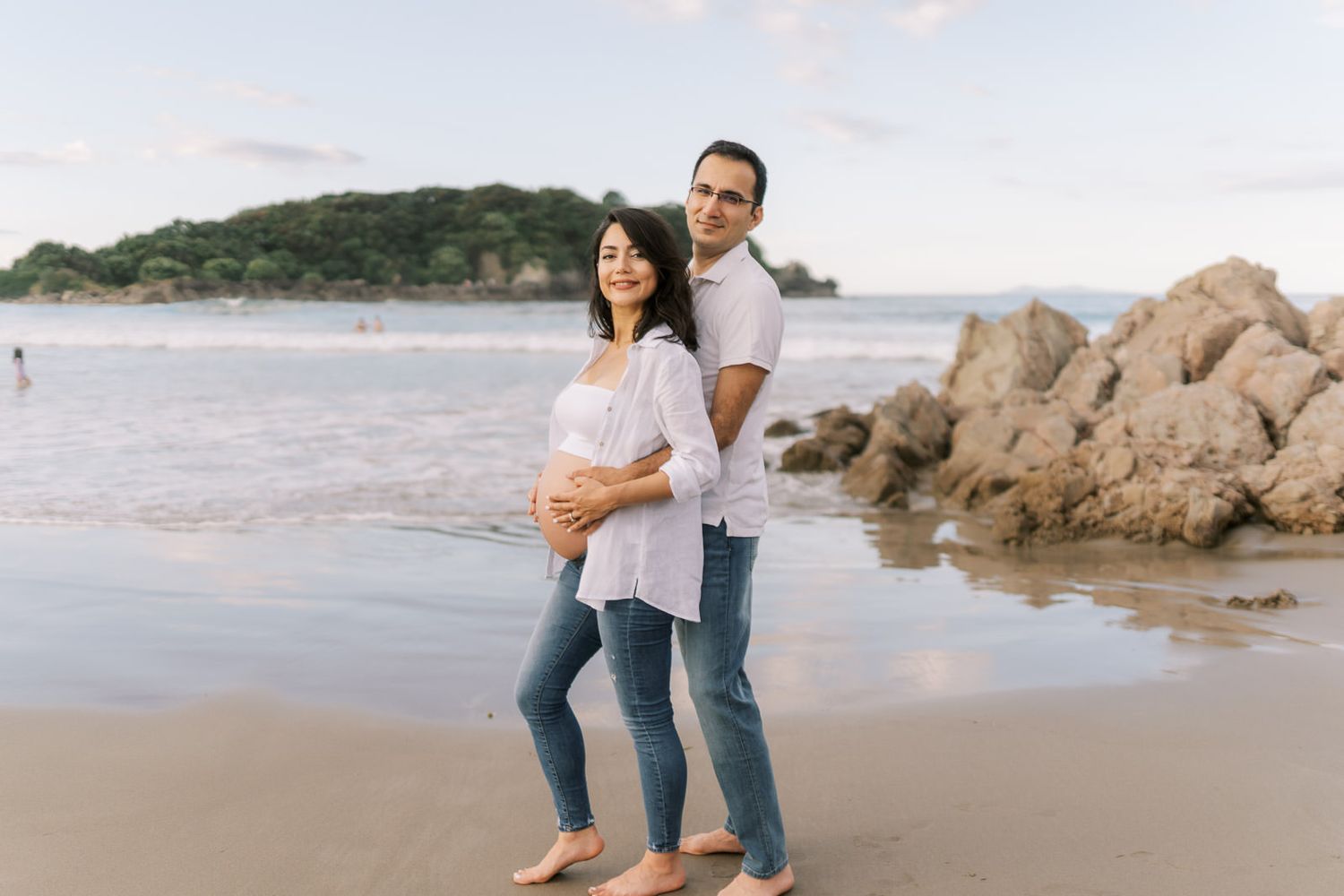 Couple posing on a beach, woman with baby bump in white top and jeans, man embracing her from behind near rocky shore.