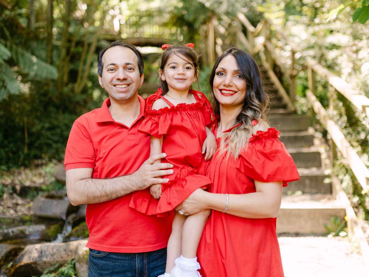 Family of three in matching red outfits smiling outdoors near wooden stairs surrounded by lush green trees.
