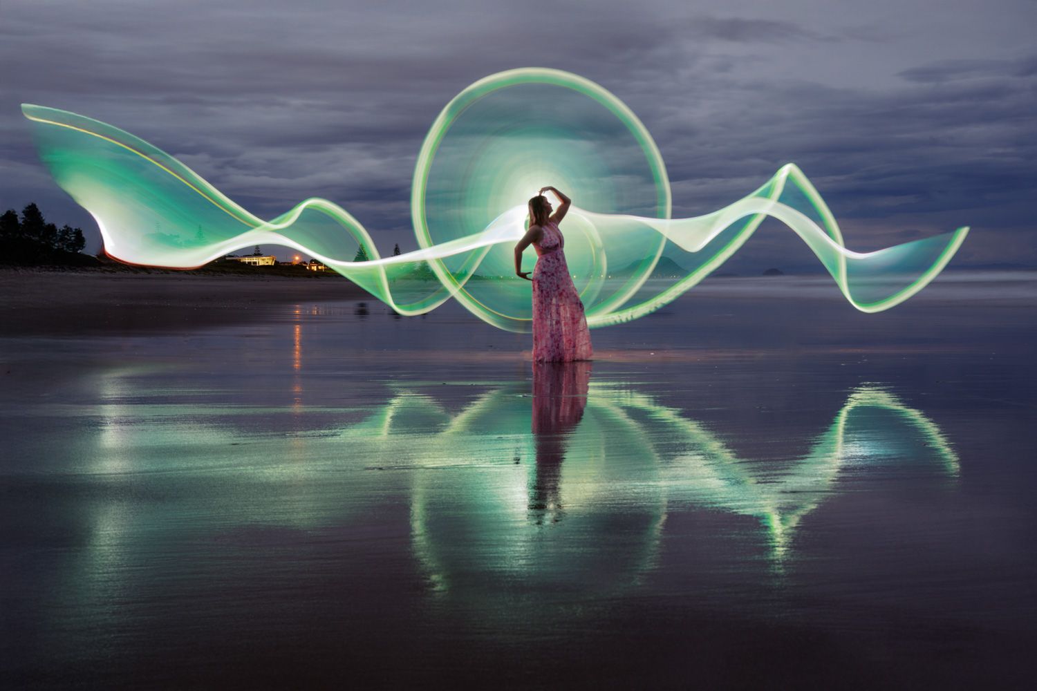 Woman in pink dress surrounded by glowing green light painting waves on a reflective beach at dusk.