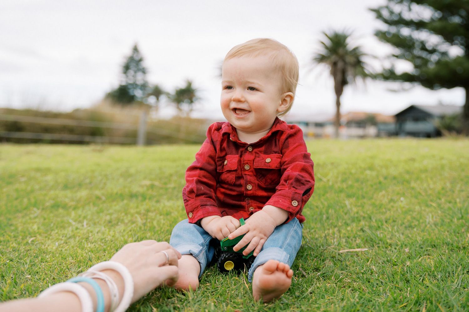 Happy baby boy in red plaid shirt sitting on green grass, smiling and holding a toy outdoors.