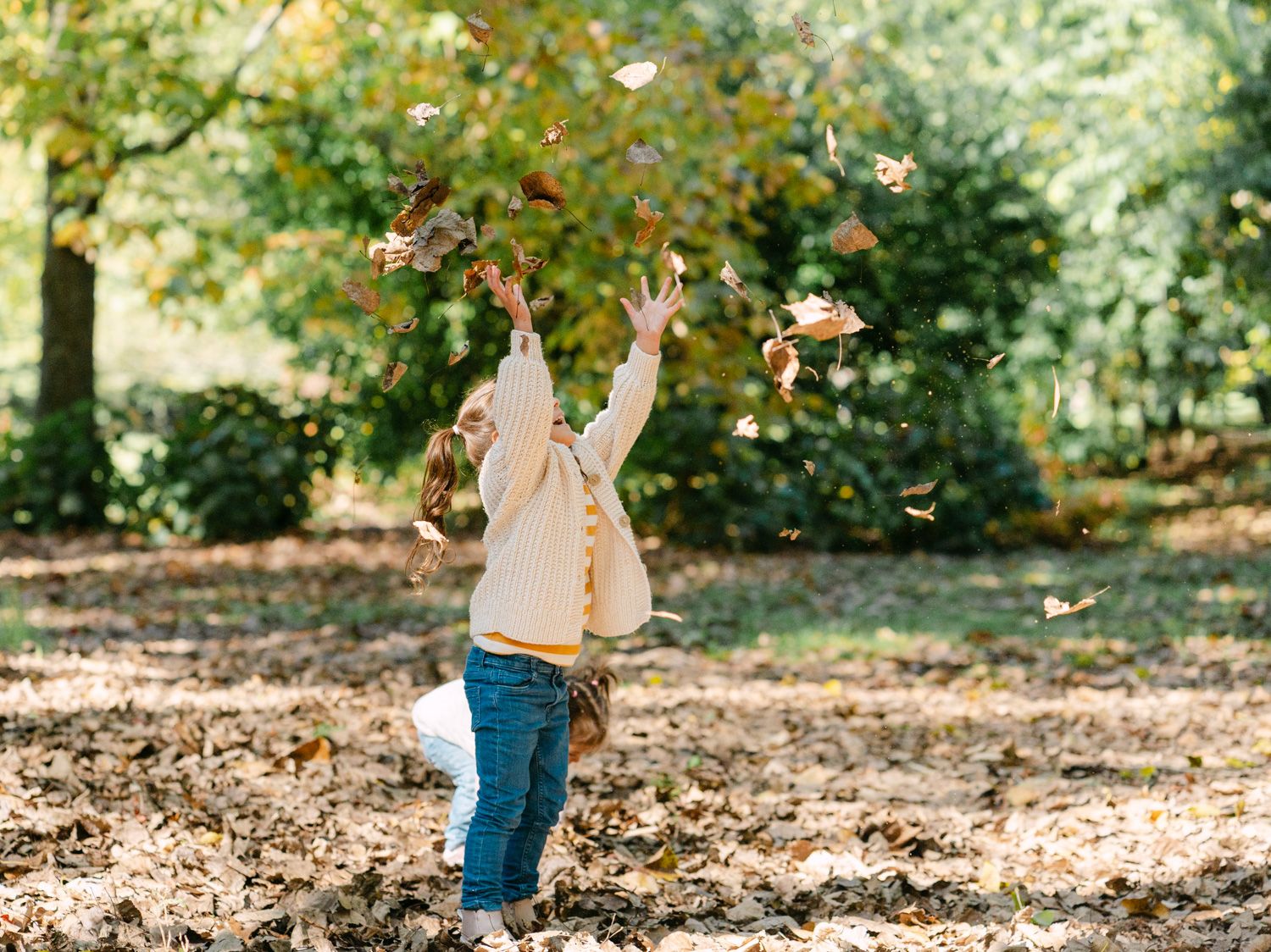 A young child joyfully tosses autumn leaves into the air in a park surrounded by colorful fall foliage.