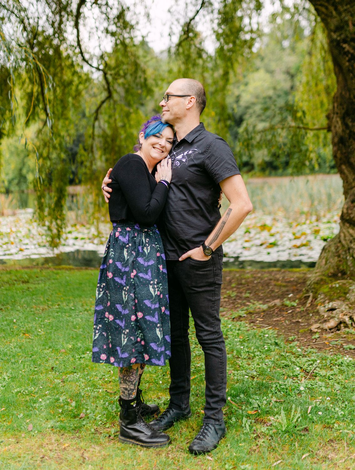 Couple embracing outdoors near a willow tree, woman with blue hair in floral skirt, man in black outfit.