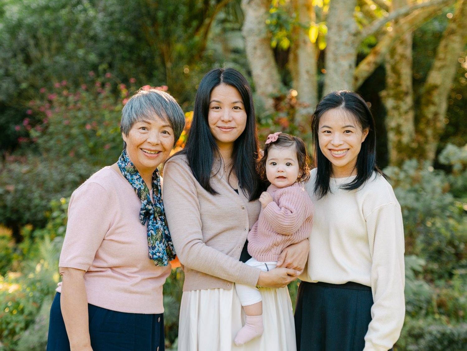 Three generations of women pose outdoors, with the middle woman holding a baby girl, surrounded by lush greenery.