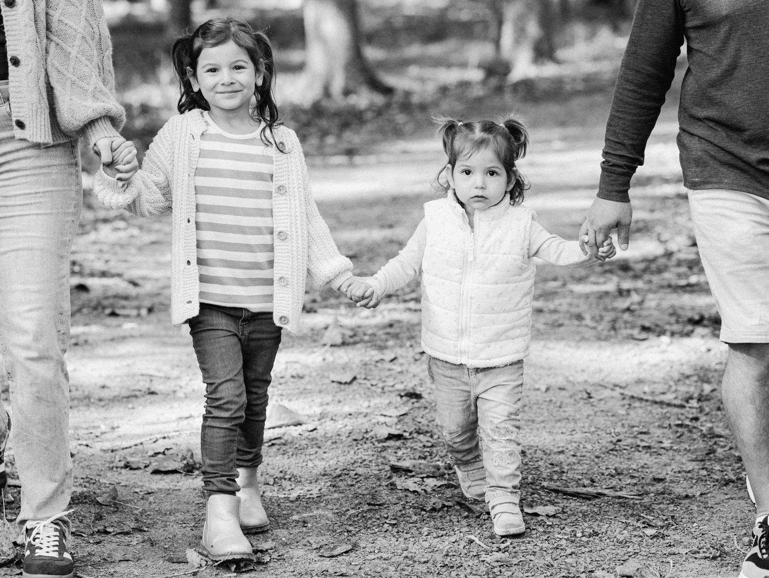 Black and white photo of two young girls holding hands with parents while walking outdoors in a park setting.