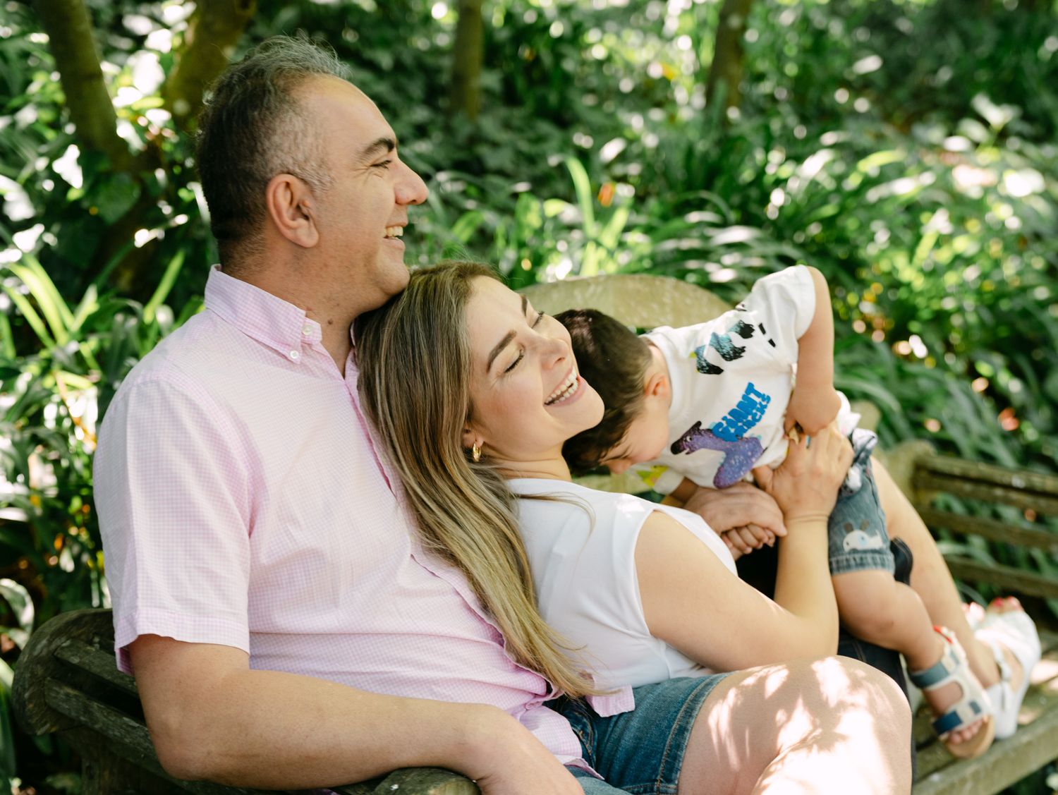 Family sitting on a bench outdoors, laughing together in a lush green garden setting.