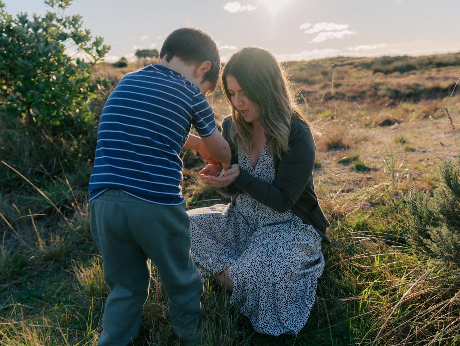 A mother kneels in a golden field, sharing a tender moment with her young son at sunset.