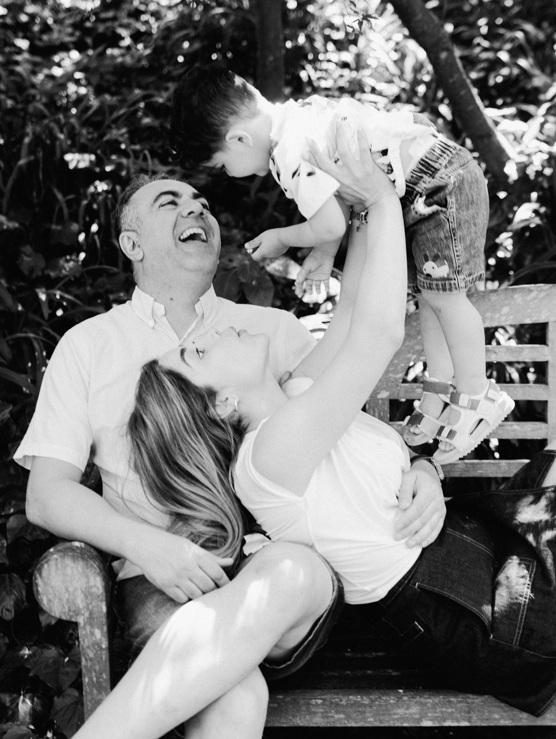 Black and white photo of a laughing family on a bench, mother holding toddler up while father smiles joyfully outdoors.