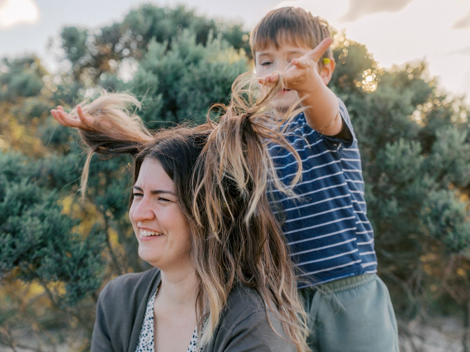 A smiling woman outdoors with a young boy sitting on her shoulders playfully pulling her hair.