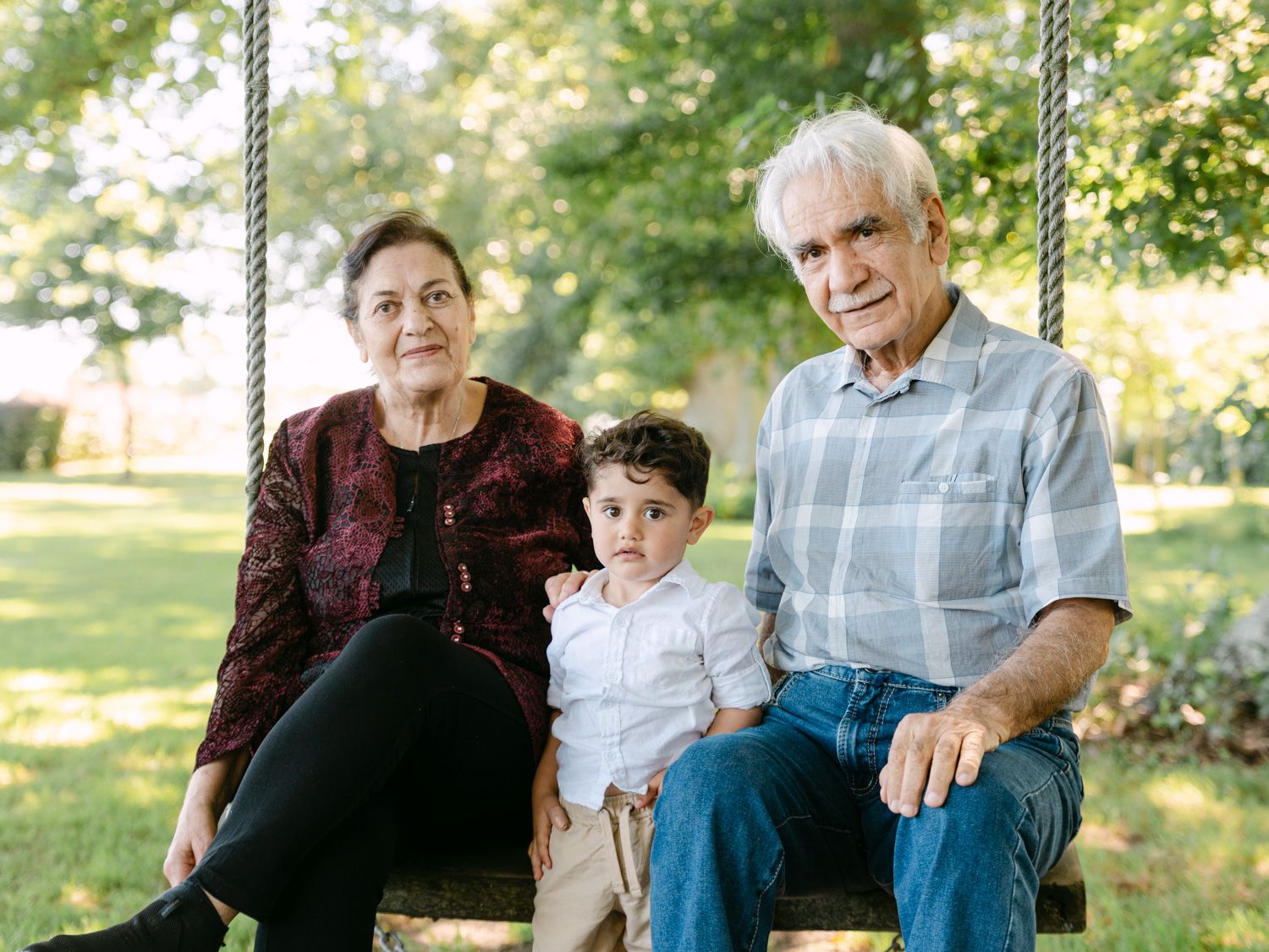 Grandparents and young grandchild sitting together on a wooden swing outdoors in a lush green garden.