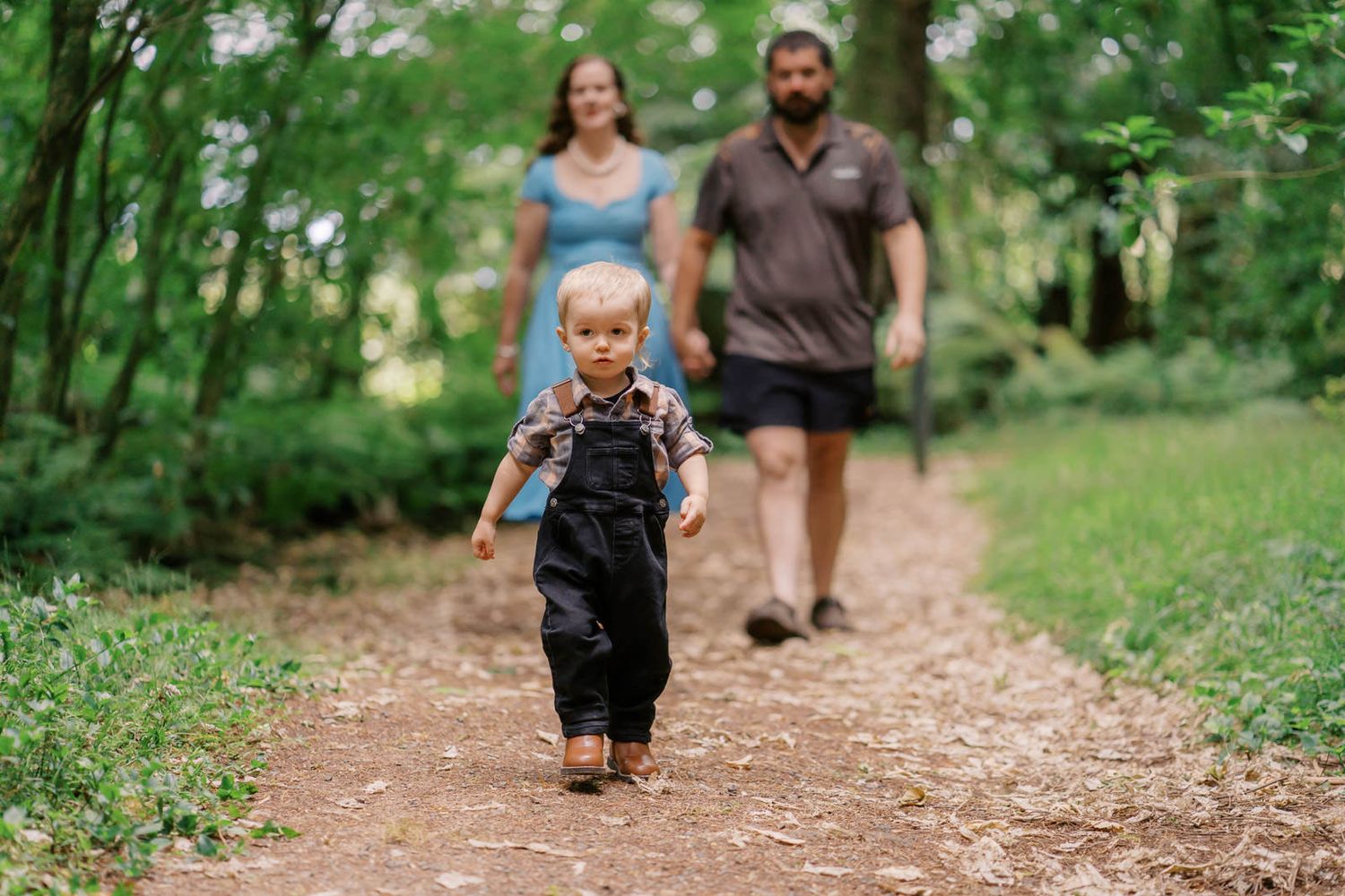Toddler boy running ahead of parents on a forest path surrounded by lush green trees.