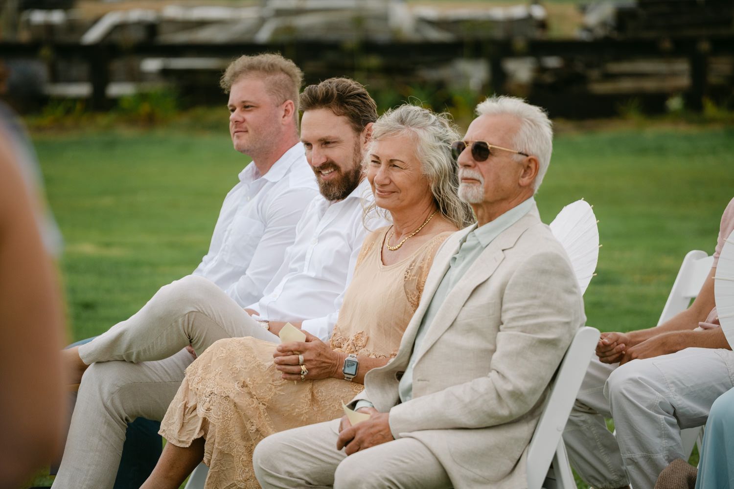 Guests seated outdoors at a wedding ceremony, smiling and watching the event on a sunny day.