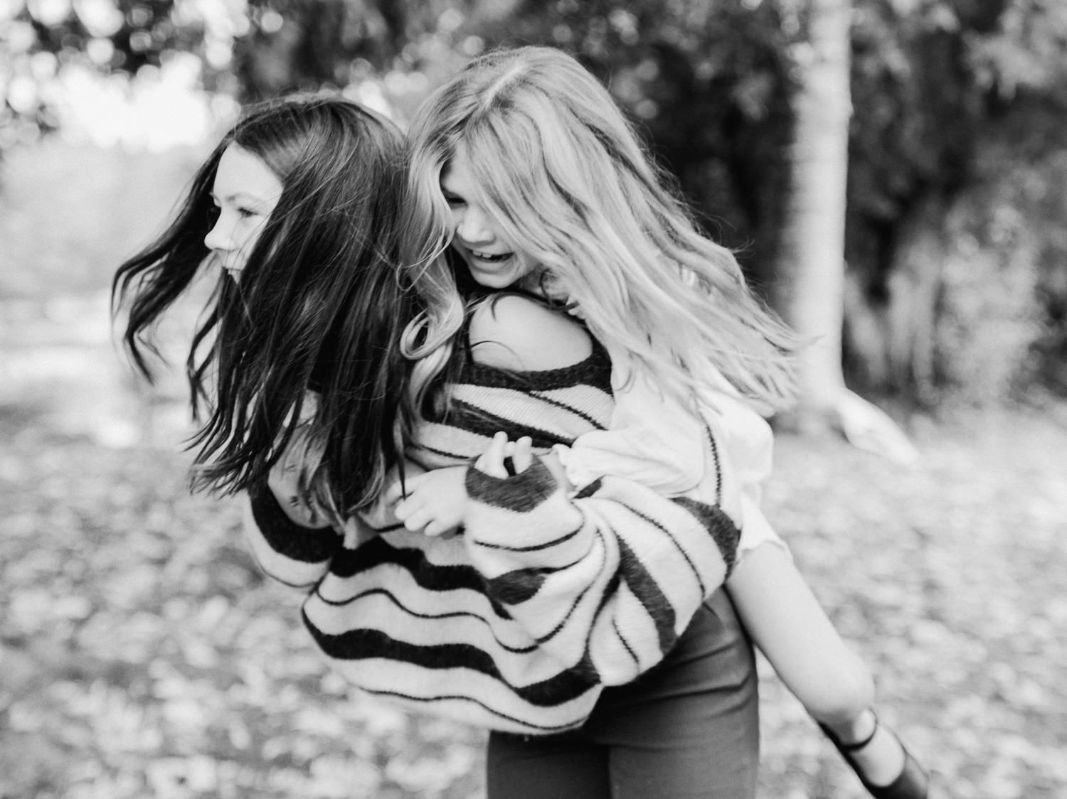 Two girls laughing outdoors, one giving the other a piggyback ride in a black and white photo.
