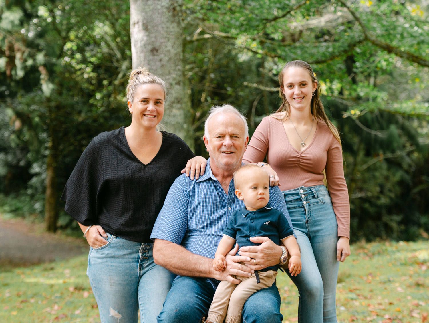 Family of three adults and a baby posing outdoors in a lush green park setting for a portrait photo.