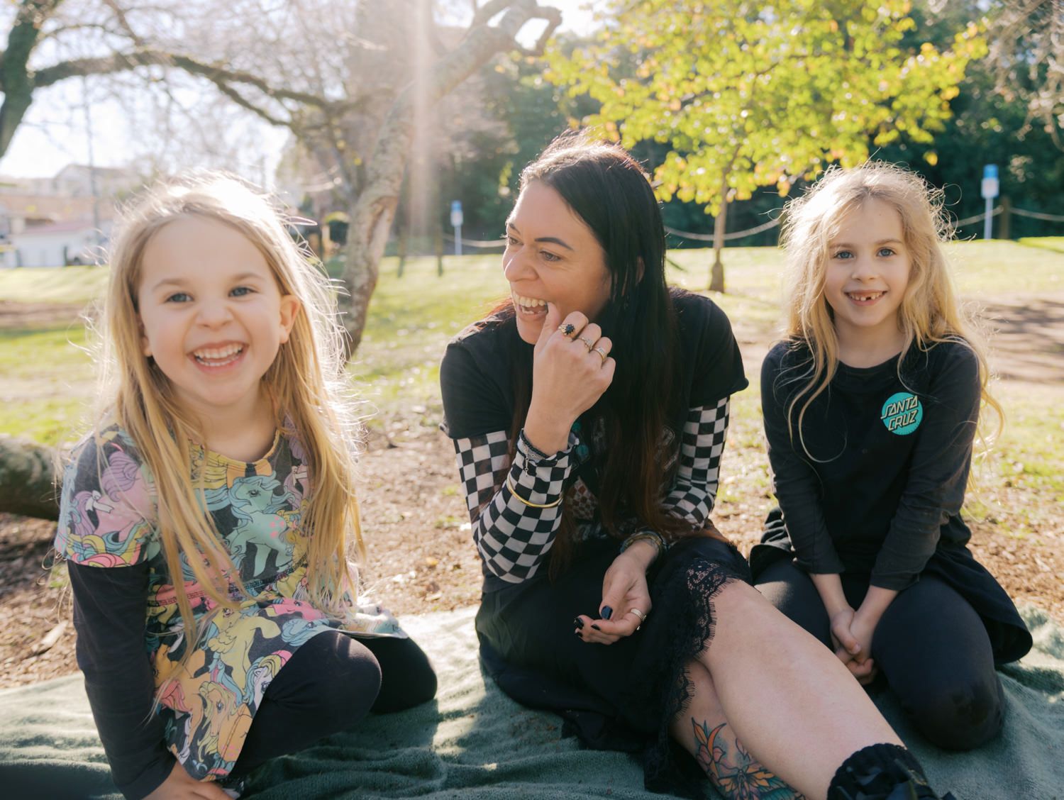 Woman and two blonde girls smiling and sitting together on a blanket in a sunny park.