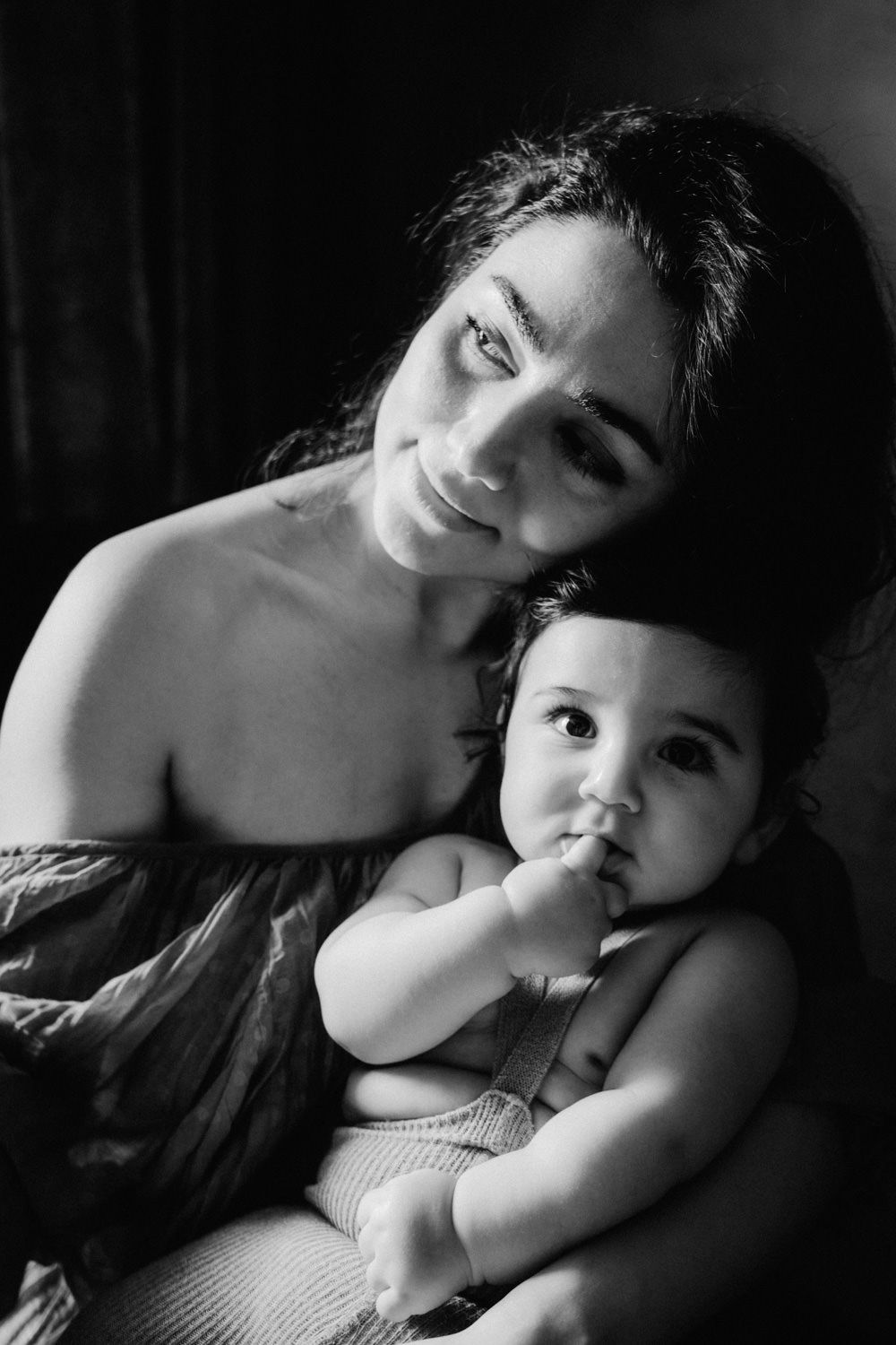 Black and white portrait of a smiling mother tenderly holding her curious infant baby with finger in mouth.