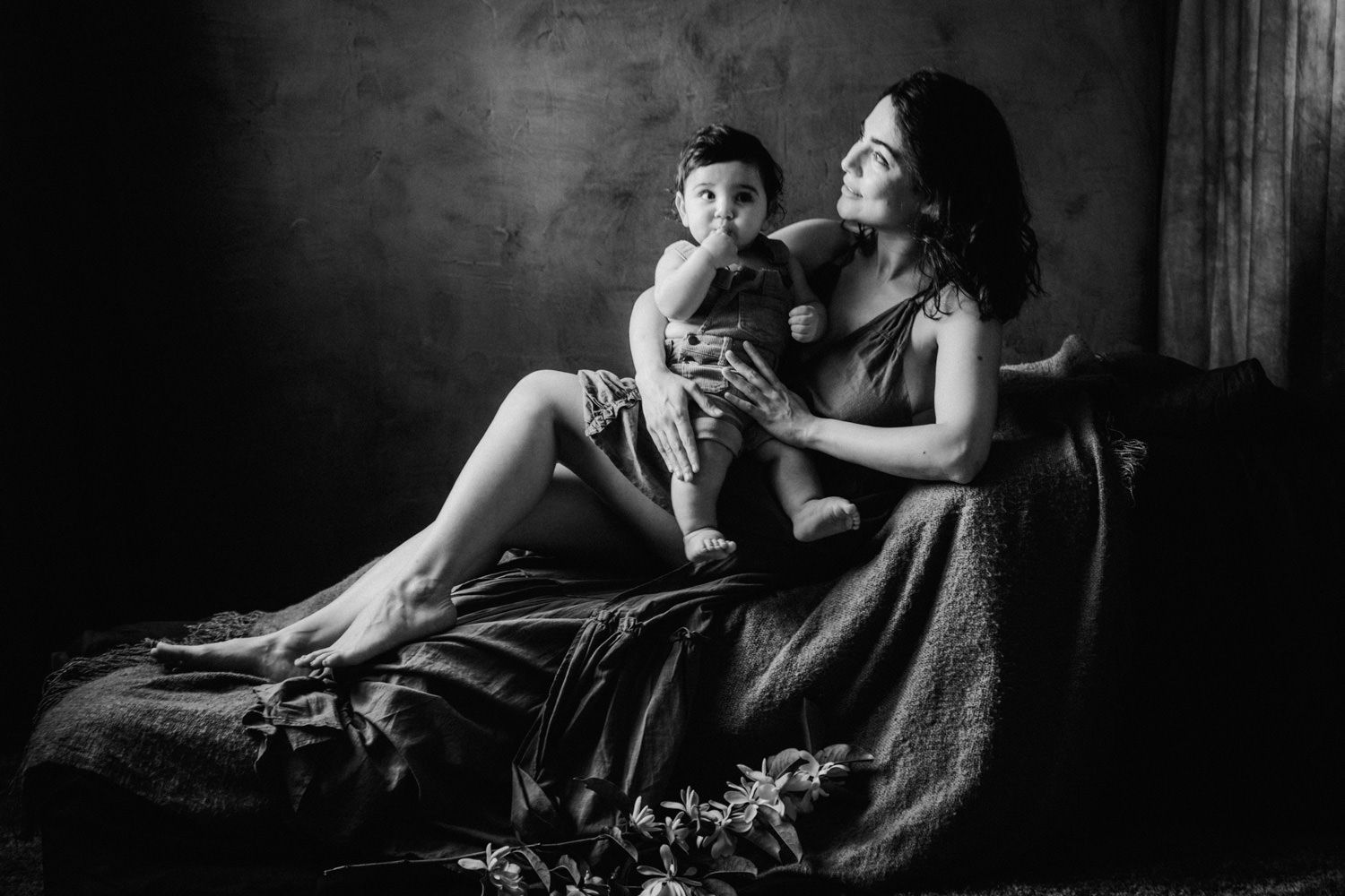 Black and white studio portrait of a mother in elegant drape holding her baby on a vintage chaise lounge.