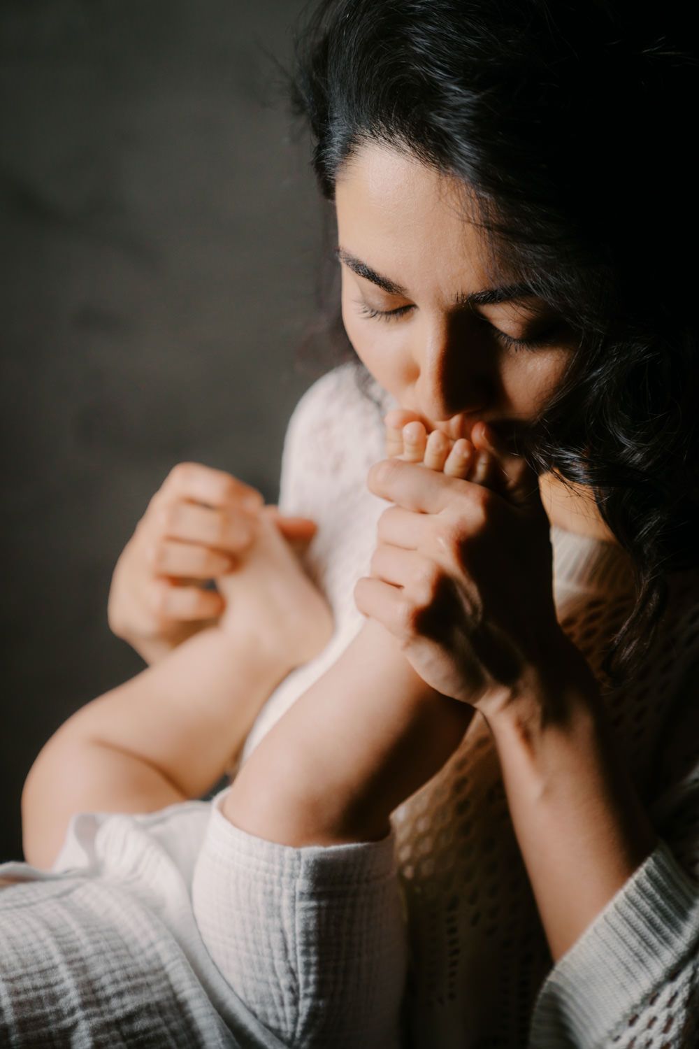 A tender mother gently kisses her newborn baby's tiny feet in warm, natural light.